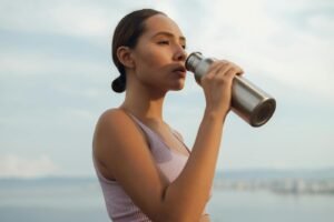 A woman drinks from a metal bottle on a sunny day by the beach, wearing a sports bra.